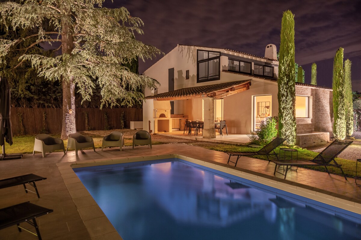 The house, set against a nighttime backdrop, is surrounded by well-maintained greenery. A serene pool reflects the evening sky, while the terrace features a dining area and a barbecue. Comfortable lounge chairs are positioned alongside the pool, inviting relaxation.