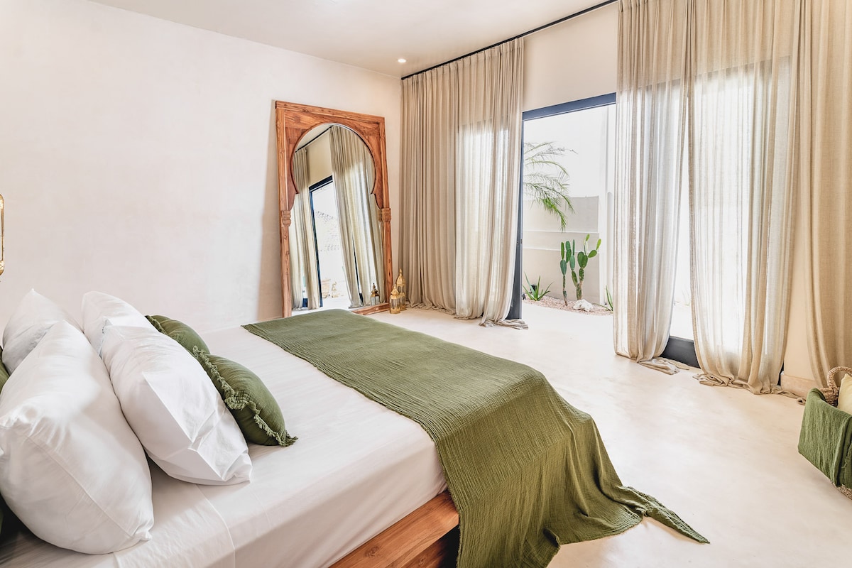 A serene bedroom features a spacious bed adorned with crisp white pillows and a textured green throw. Natural light filters in through sheer curtains, illuminating the room's minimalist design. A large mirror reflects the calming ambiance, while potted plants are visible beyond the window.