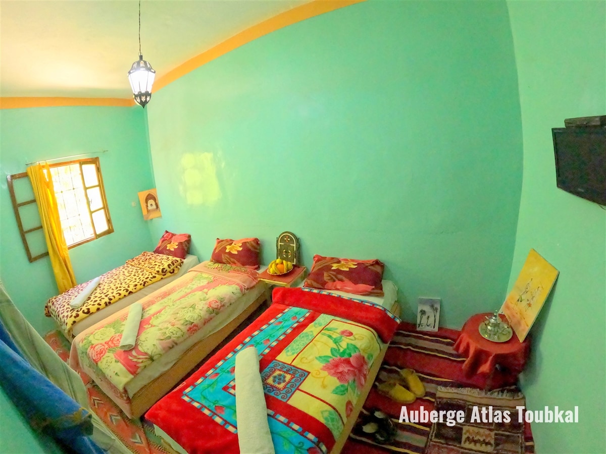 A brightly colored room features three spacious beds adorned with patterned bedspreads. Light green walls create an airy atmosphere, while a window allows natural light to filter in. A small table with decorative items is positioned next to one of the beds.