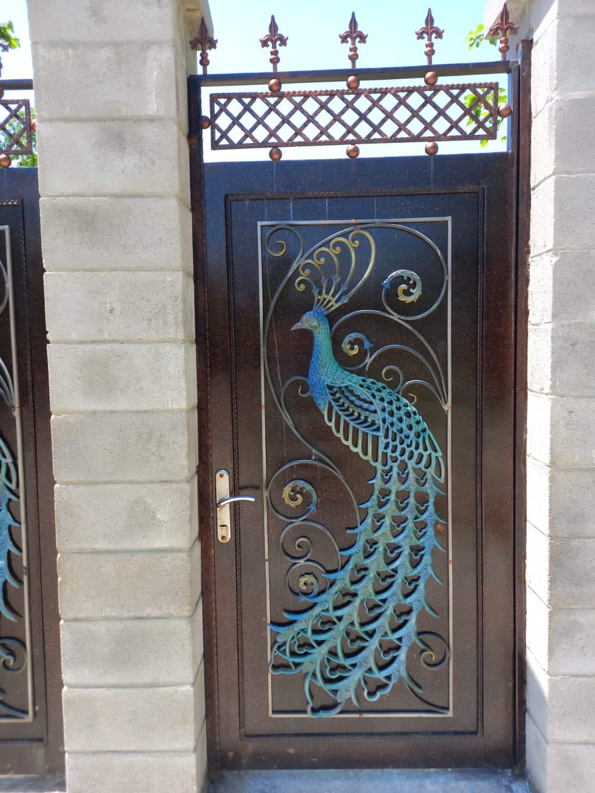 An intricately designed metal gate features a peacock motif, showcasing detailed patterns and vibrant colors. The gate is framed by sturdy stone walls, with decorative latticework above, allowing glimpses of greenery beyond.