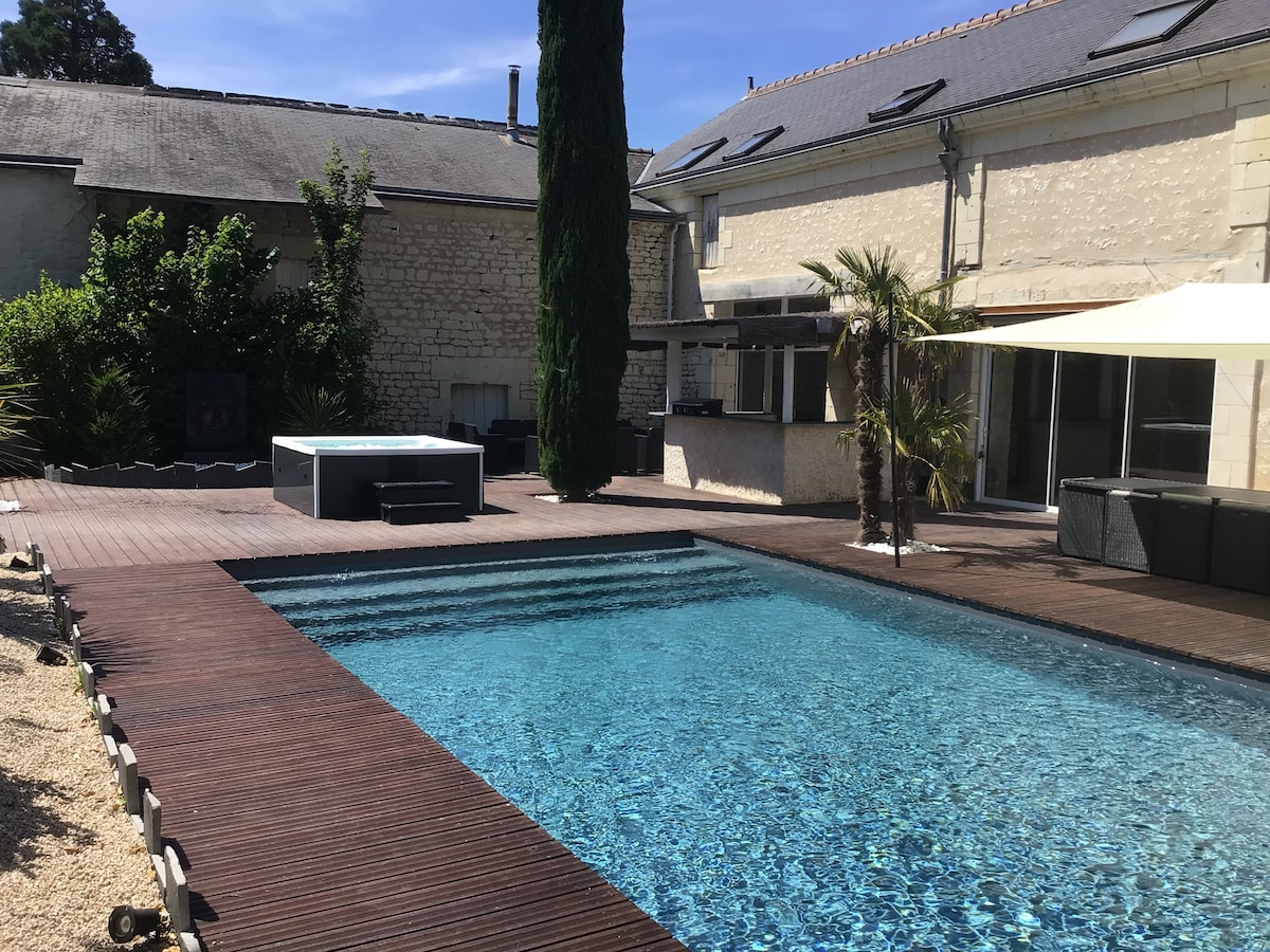 A tranquil outdoor area features a heated swimming pool surrounded by a wooden deck. A jacuzzi is positioned near the pool. Lush greenery and a shaded seating area enhance the inviting atmosphere, with a barbecue grill visible under the shade of an umbrella.
