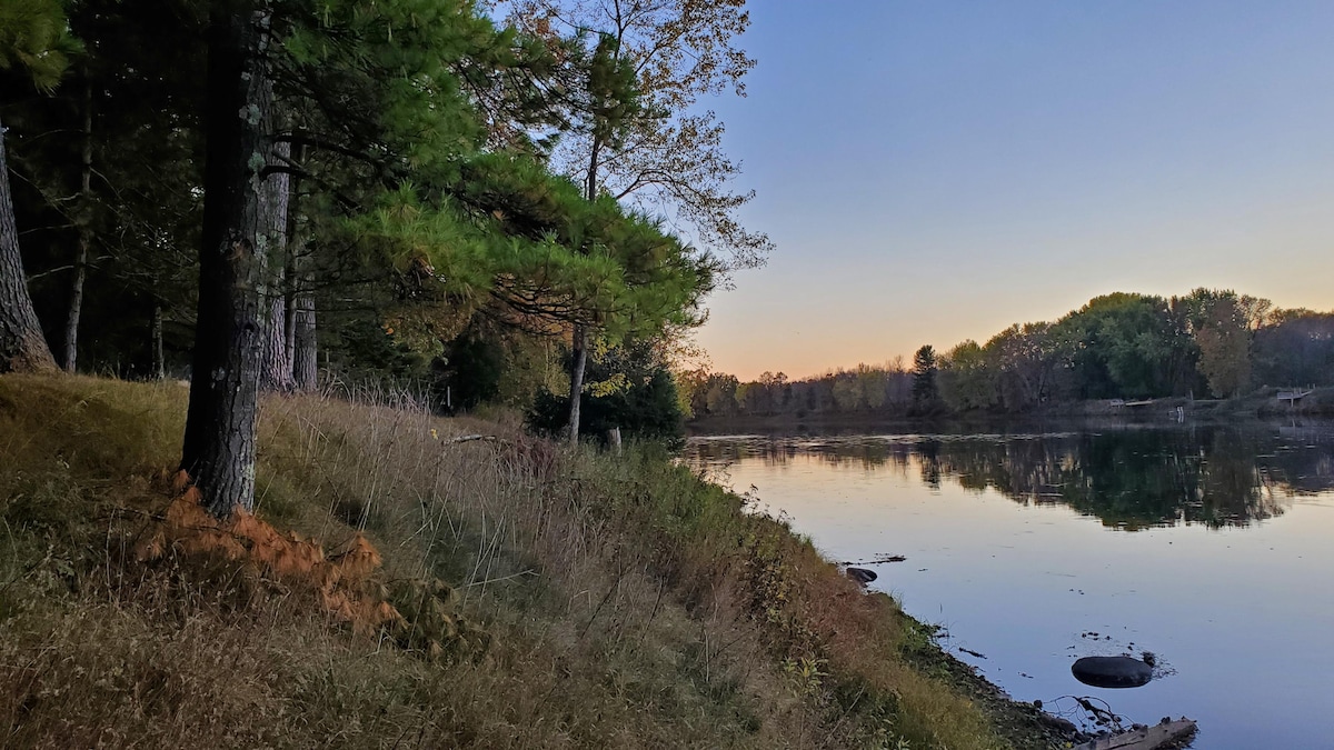 A serene view of the Menominee River is captured at dusk, surrounded by lush greenery and tall pines. The gentle slope leads toward the water's edge, reflecting the soft hues of the evening sky and offering a peaceful atmosphere for outdoor activities.
