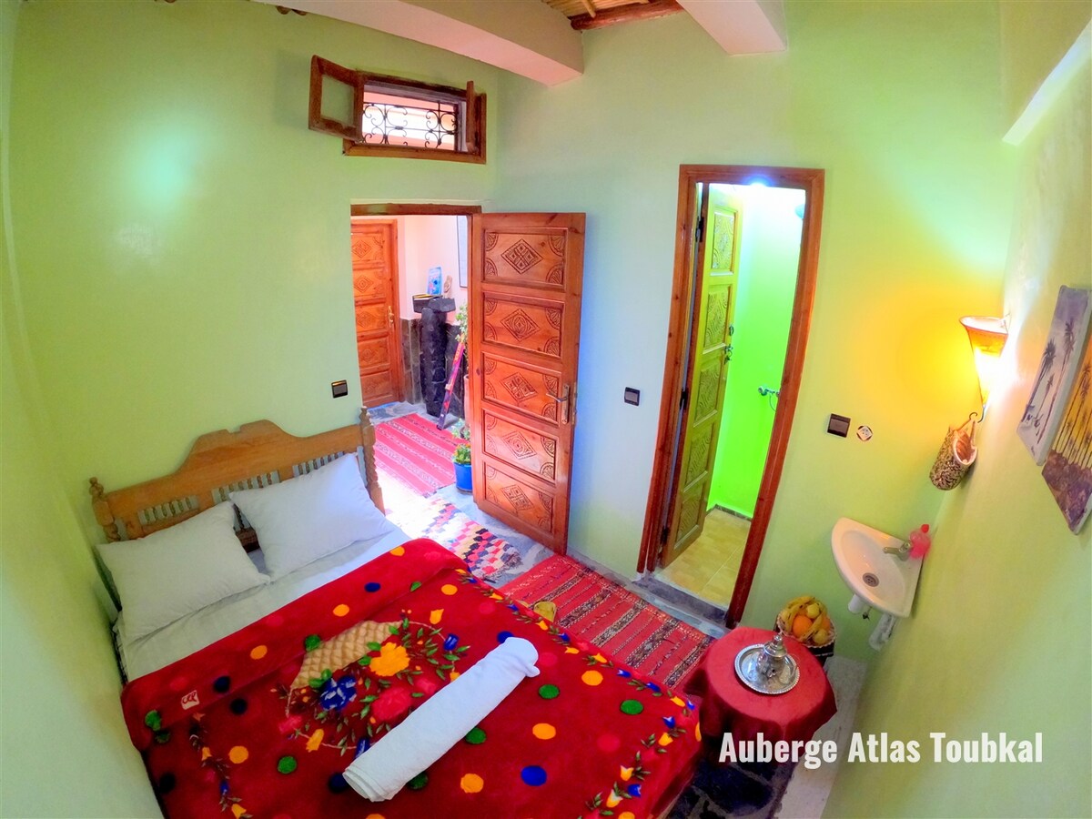 A compact sleeping area features a spacious bed adorned with a colorful blanket. Wooden doors lead to two separate areas, and a small washbasin is visible near the entrance. Decorative elements and vibrant textiles add character to the room's green walls.
