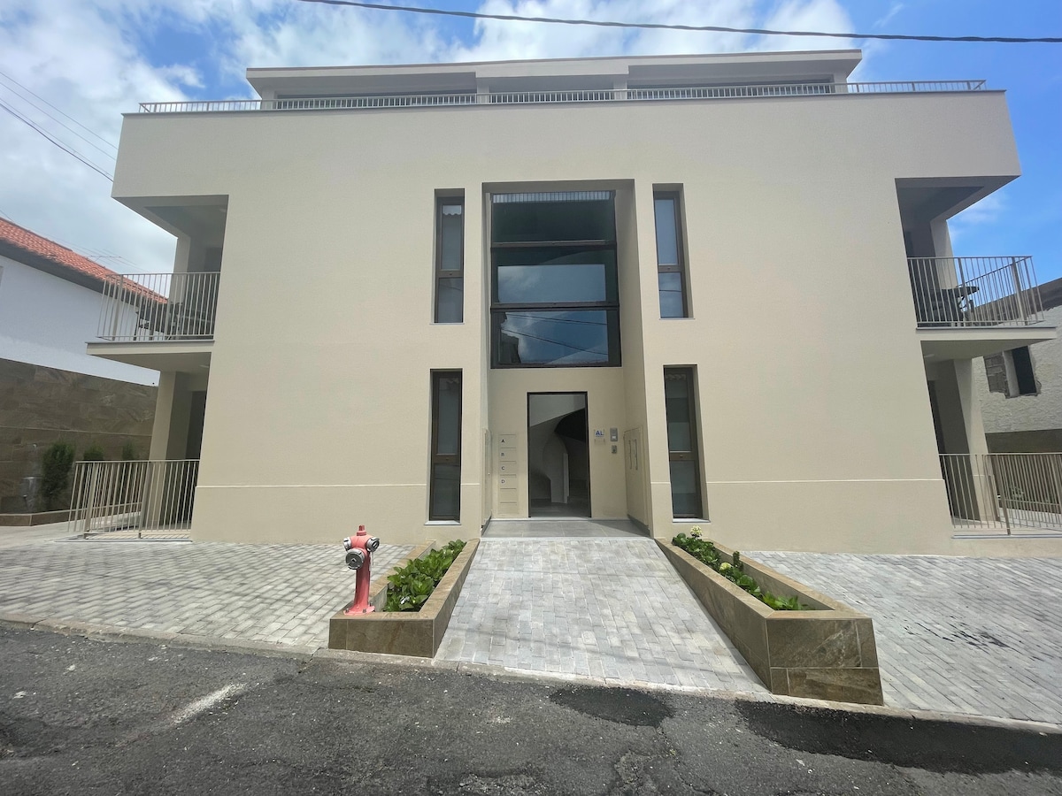 A modern three-story building is visible, featuring a minimalist facade in neutral tones. Large windows allow natural light to enter. The entrance is marked by a central doorway with a sleek design, flanked by small garden areas and a paved path leading to the entrance.