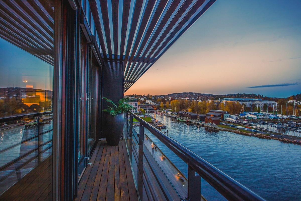 A balcony with wooden decking offers a panoramic view of the water at sunset. Horizontal slats create a pattern against the sky, while a large plant is positioned in a corner near a glass railing. The scene captures a serene waterfront with boats and distant hills.