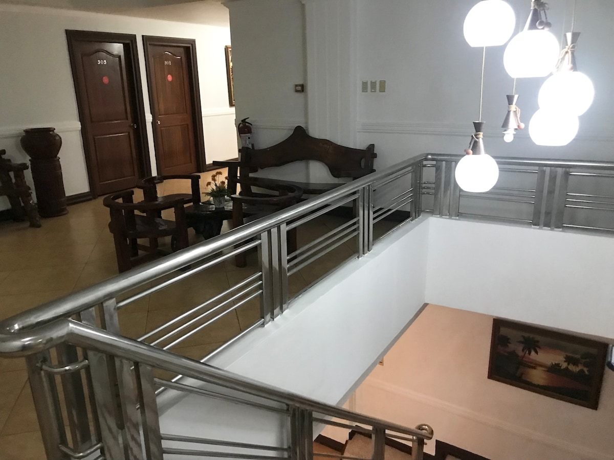 A well-lit staircase is framed by polished stainless steel railings. Wooden doors are visible along the corridor. A seating area with a circular table and chairs can be seen beside the staircase, accompanied by decorative plants.