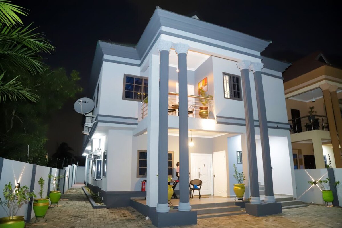 The exterior of a two-story house features tall columns and a mix of gray and white walls. Soft lighting highlights the entrance, where potted plants are positioned. A patio area with seating is visible, surrounded by a well-maintained compound with greenery.