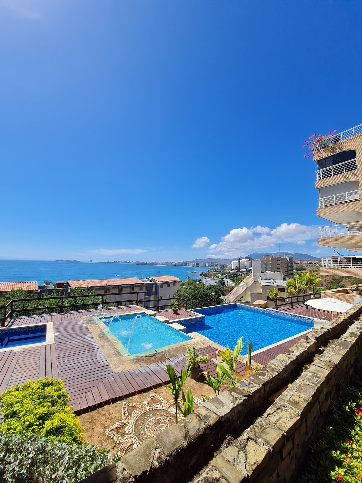 An outdoor area features a large blue swimming pool and a children's pool surrounded by a wooden deck. Lush greenery is visible, along with a panoramic view of the bay under a bright blue sky. Nearby, a building rises, adding depth to the scene.