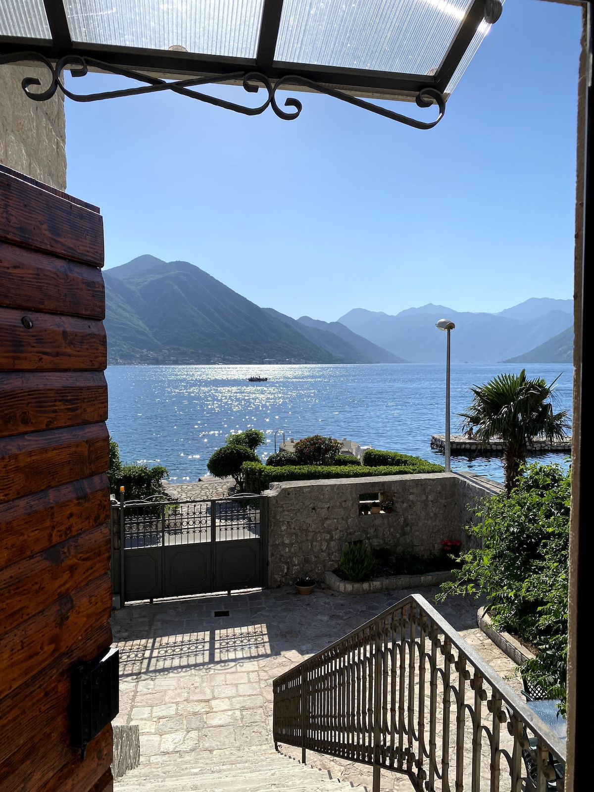 A view from an open door reveals a scenic landscape of mountains and water. The sun reflects off the tranquil lake, while stone steps lead down to a lush garden. A palm tree and greenery frame the entrance, creating an inviting outdoor space.