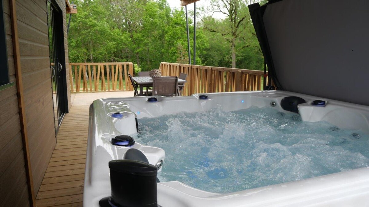 A hot tub is situated on a wooden deck, surrounded by lush greenery. The bubbling water invites relaxation. In the background, a seating area with a table and chairs is visible, enhancing the outdoor experience.