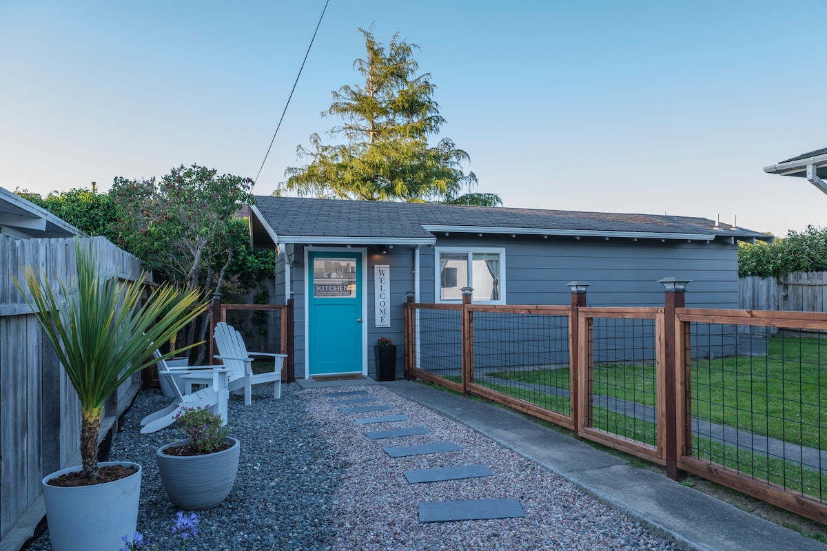 A charming entrance to the studio features a gravel pathway lined with stepping stones. Two Adirondack chairs are positioned beside a small garden area, while a fence surrounds the lush lawn, creating a private outdoor space. The main door is painted in a vibrant blue.