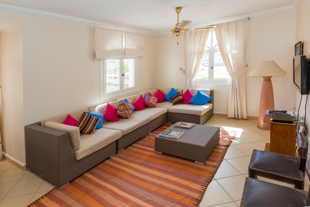 A light-filled living area features a large sectional sofa adorned with colorful cushions. A coffee table sits in the center, complemented by a patterned area rug. Natural light enters through multiple windows with sheer curtains, creating a bright and inviting space.