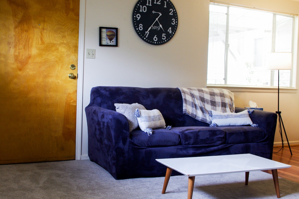 A living area is depicted, featuring a plush blue couch adorned with decorative pillows. A light-colored coffee table is positioned in front, creating a functional space. Large windows provide natural light, while a clock is mounted on the wall above. Carpeted flooring enhances the comfort.