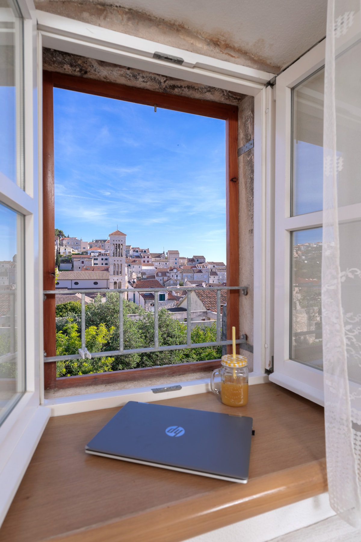 A window offers a view of Hvar's charming rooftops and the bell tower of St. Stephen's Cathedral. A small table with a laptop and a beverage sits on a wooden ledge, illuminated by natural light filtering through the open windows.
