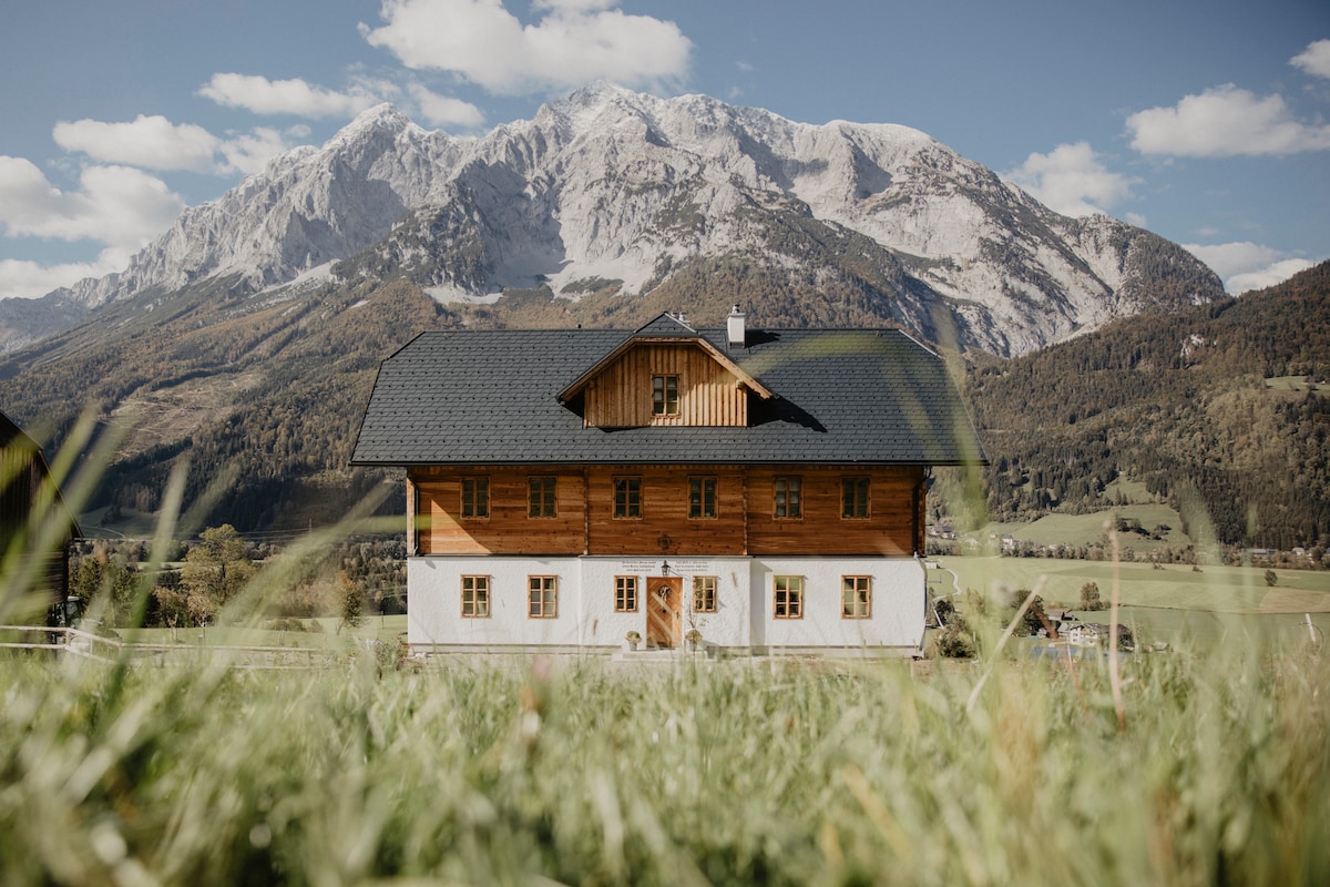 A newly built farmhouse is set against a backdrop of majestic mountains, surrounded by lush greenery. The structure features a combination of wood and white plaster, showcasing multiple windows that reflect the natural light and scenic landscape.