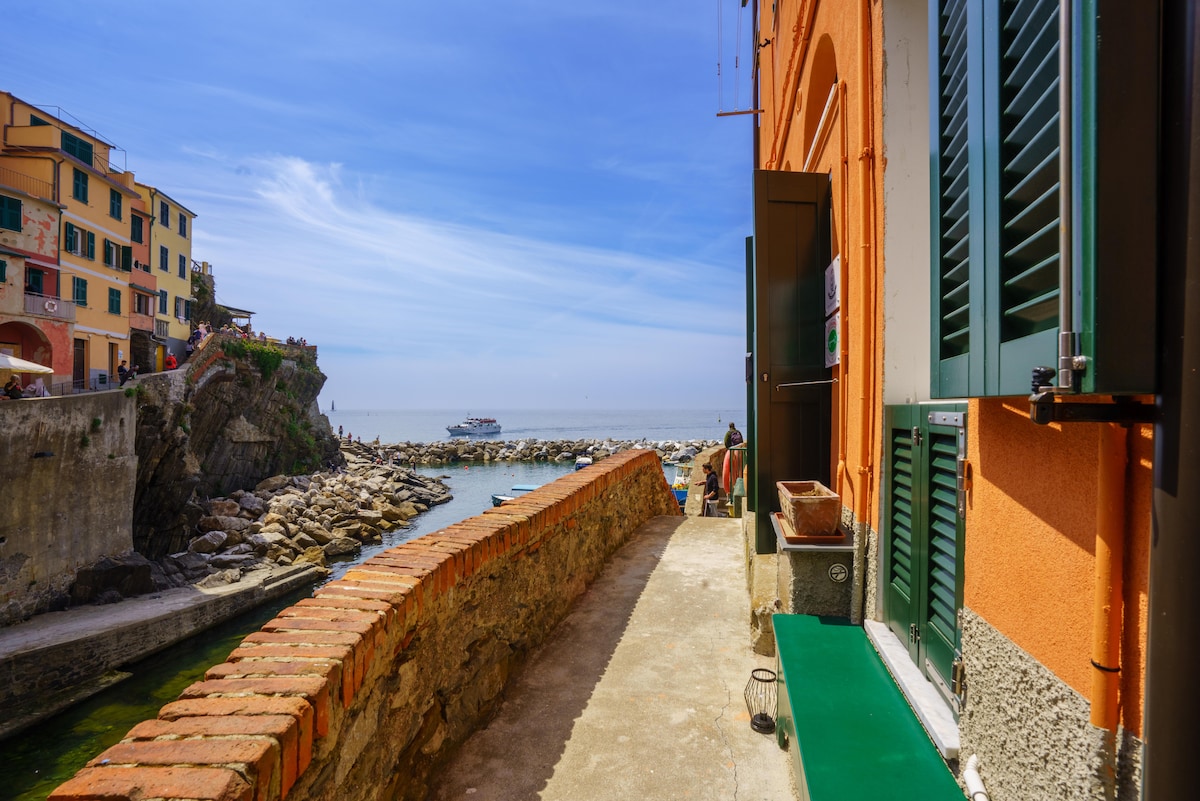 A scenic view captures the pathway along the waterfront, bordered by colorful buildings. The calm sea stretches out in the background, with a small boat visible on the horizon. The pathway features a stone border with a smooth, paved surface.