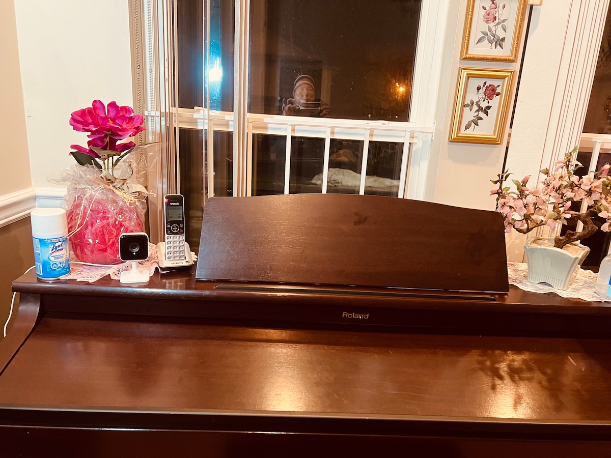 A polished wooden piano is positioned against a window, reflecting soft indoor lighting. Decorative items include a pink flower arrangement, framed art on the wall, and a small vase with flowers. A cordless phone and a container of cleaning supplies are visible on the piano's surface.