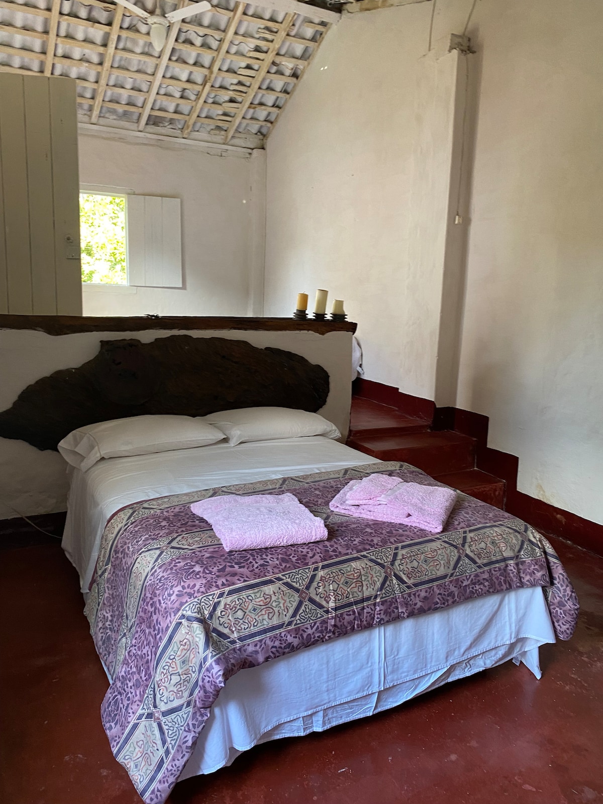 A cozy sleeping area features a spacious bed adorned with a patterned blanket and soft towels. Natural light enters through a small window, illuminating the rustic decor. Warm tones and simple furnishings create a calm and inviting space.