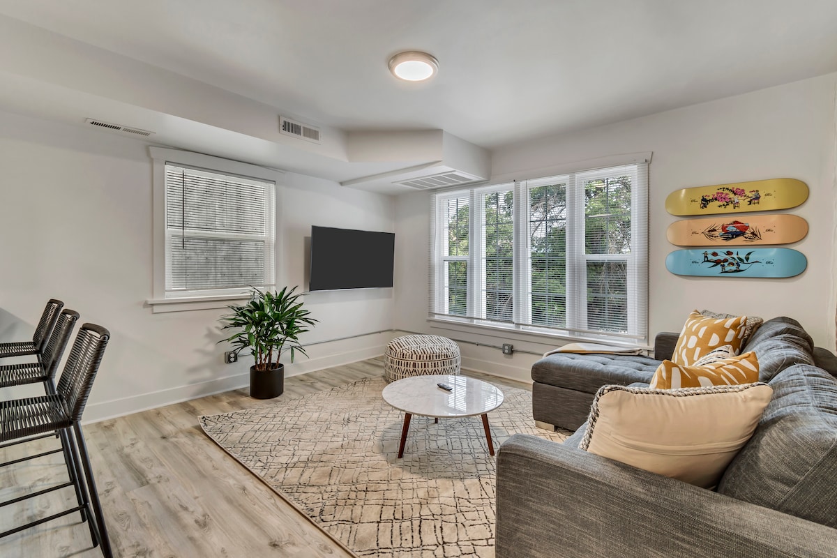 A bright living area is featured, showcasing a spacious gray sectional sofa and a round coffee table atop a textured area rug. Large windows allow natural light to fill the space, while colorful wall art adds a playful touch. Counter seating is visible in the background.