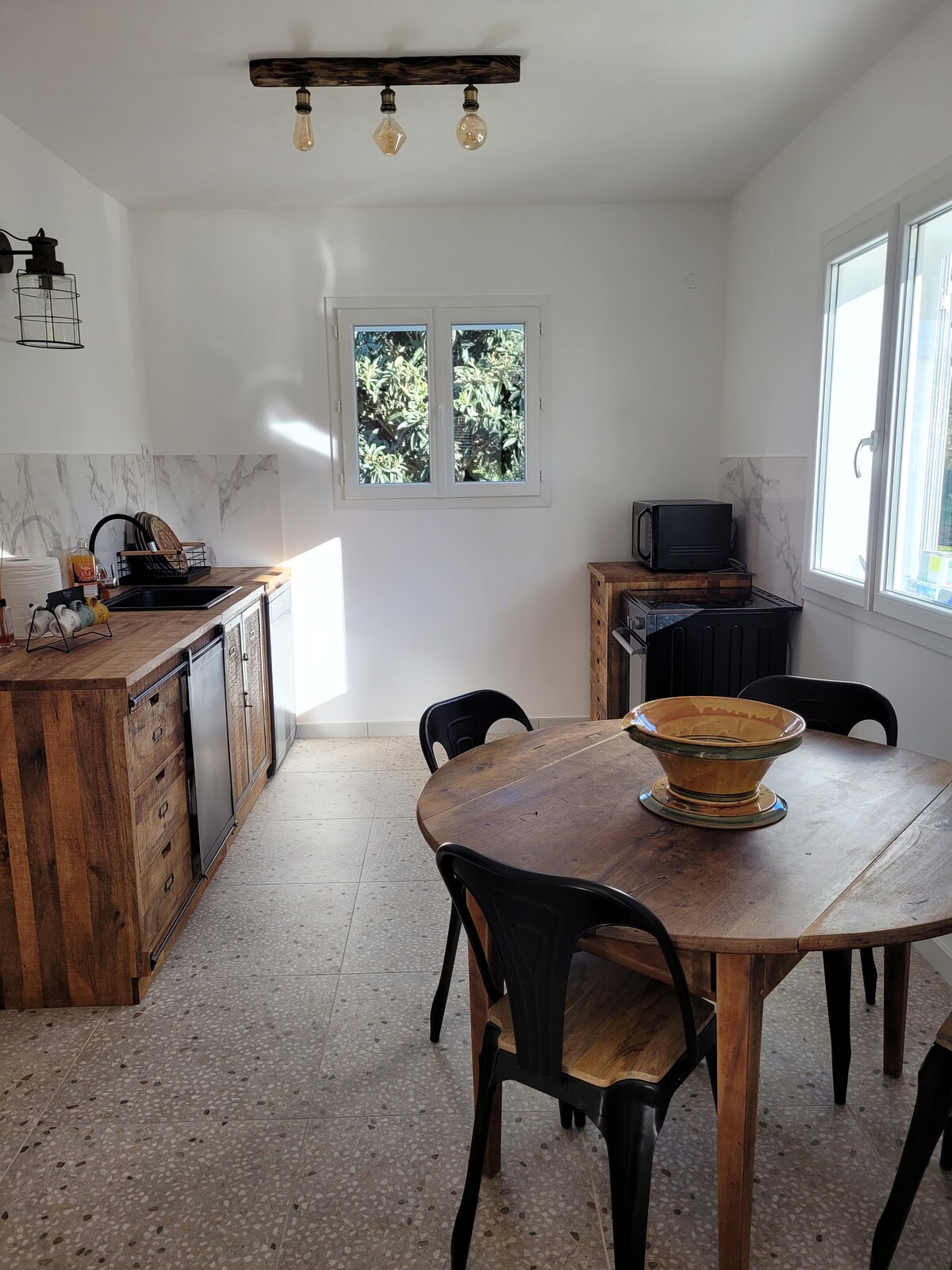 A well-lit kitchen area features a wooden dining table surrounded by six black chairs. A kitchen island with drawers and cabinets complements the space, which is equipped with modern appliances, including a microwave and a sink. Natural light streams in through two windows.