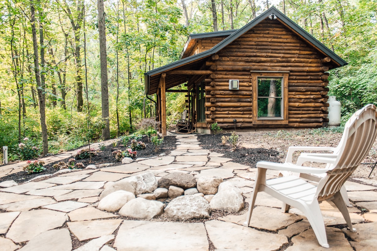 The exterior of the cabin is shown, featuring natural wooden logs and large windows. A stone pathway leads to a spacious seating area with a fire pit surrounded by large rocks. A white lawn chair is positioned near the fire pit, set among lush greenery.