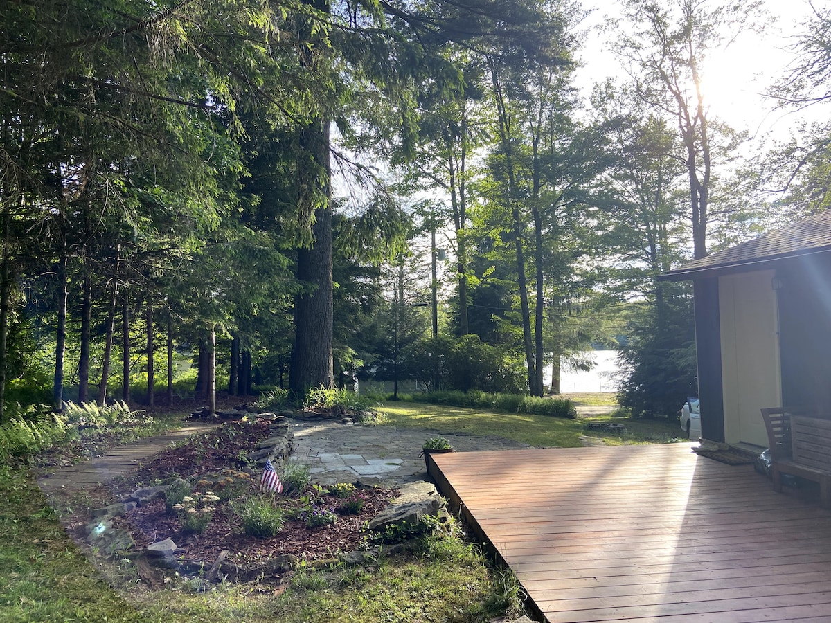 A serene outdoor space is displayed, featuring a wooden deck extending from the house. The area is surrounded by lush greenery, with tall trees and a glimpse of a lake visible in the background. Sunlight filters through the branches, creating a peaceful ambiance.