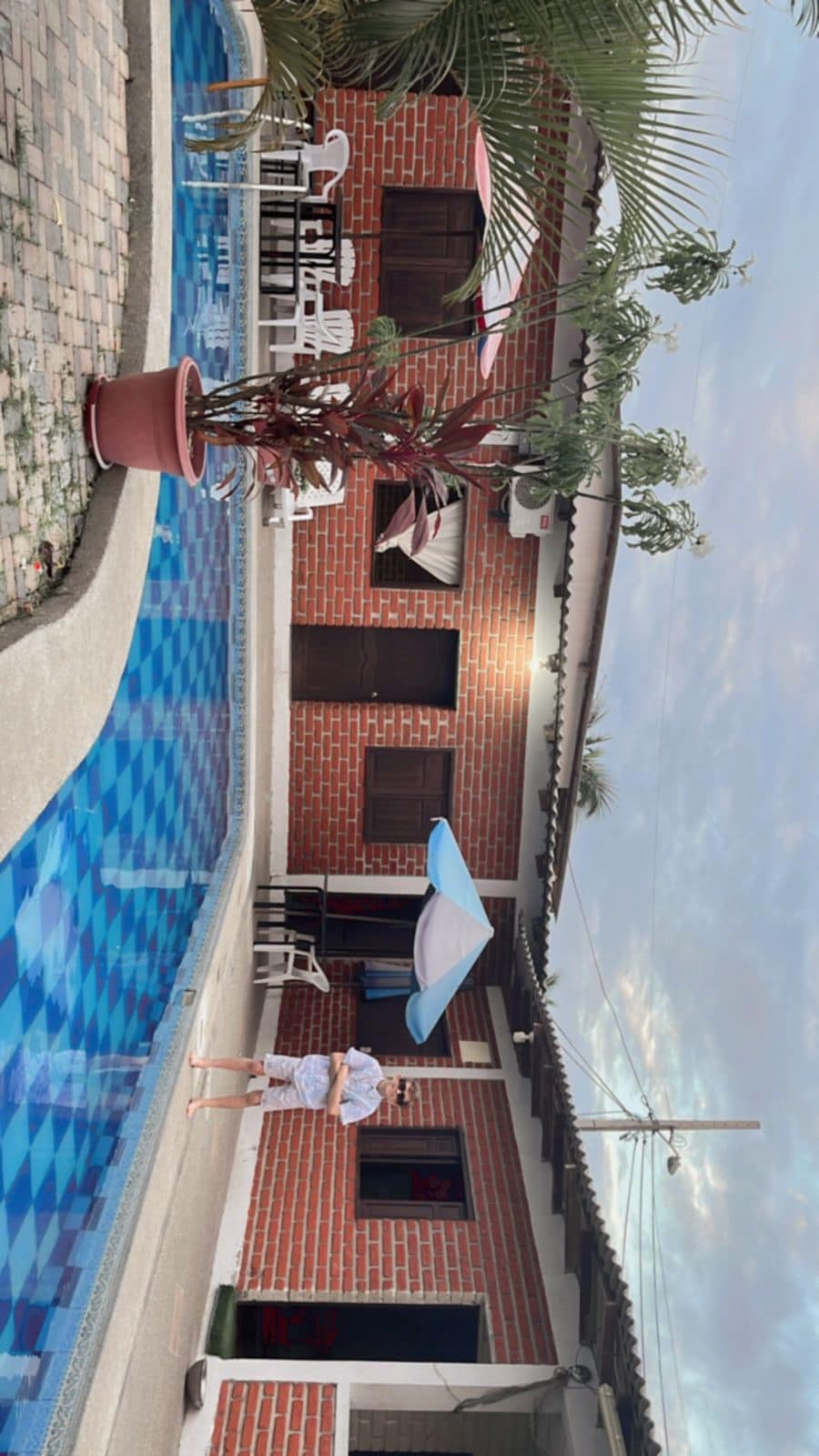 A refreshing swimming pool reflects the surrounding area, featuring a vibrant blue tile pattern. Lounging chairs position near the water, complemented by a shaded area under a blue umbrella. The entrance to a cozy brick house is visible, alongside potted plants adding greenery.