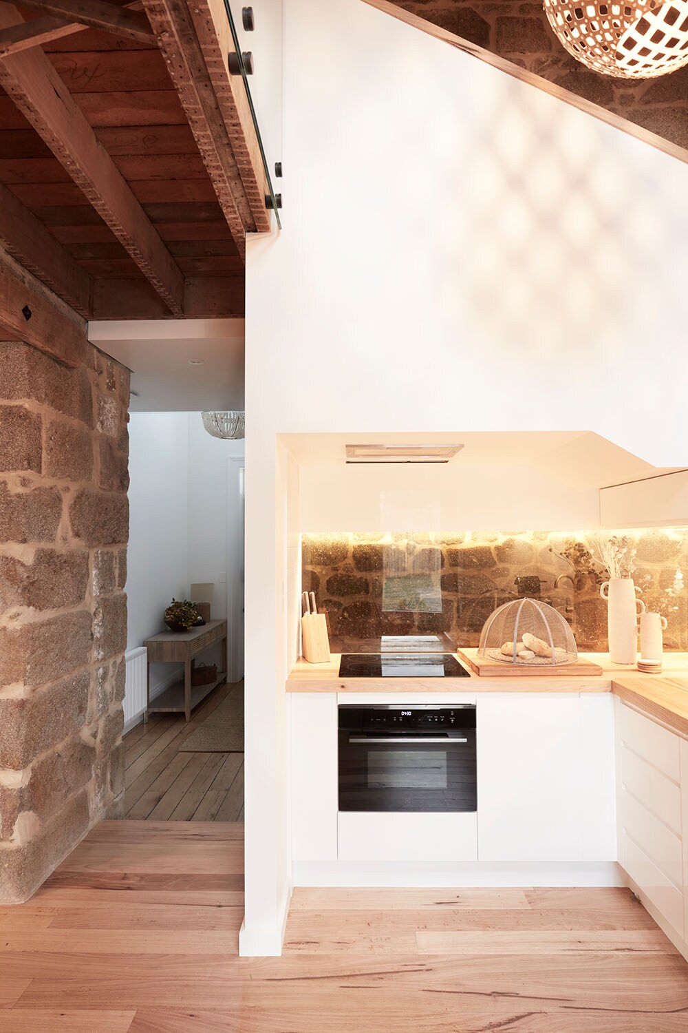 A modern kitchen area features sleek white cabinetry and a wooden countertop, complemented by warm lighting. The original granite stone wall adds character, while contemporary appliances are neatly integrated. An open layout connects the kitchen to the hallway, enhancing the sense of space.