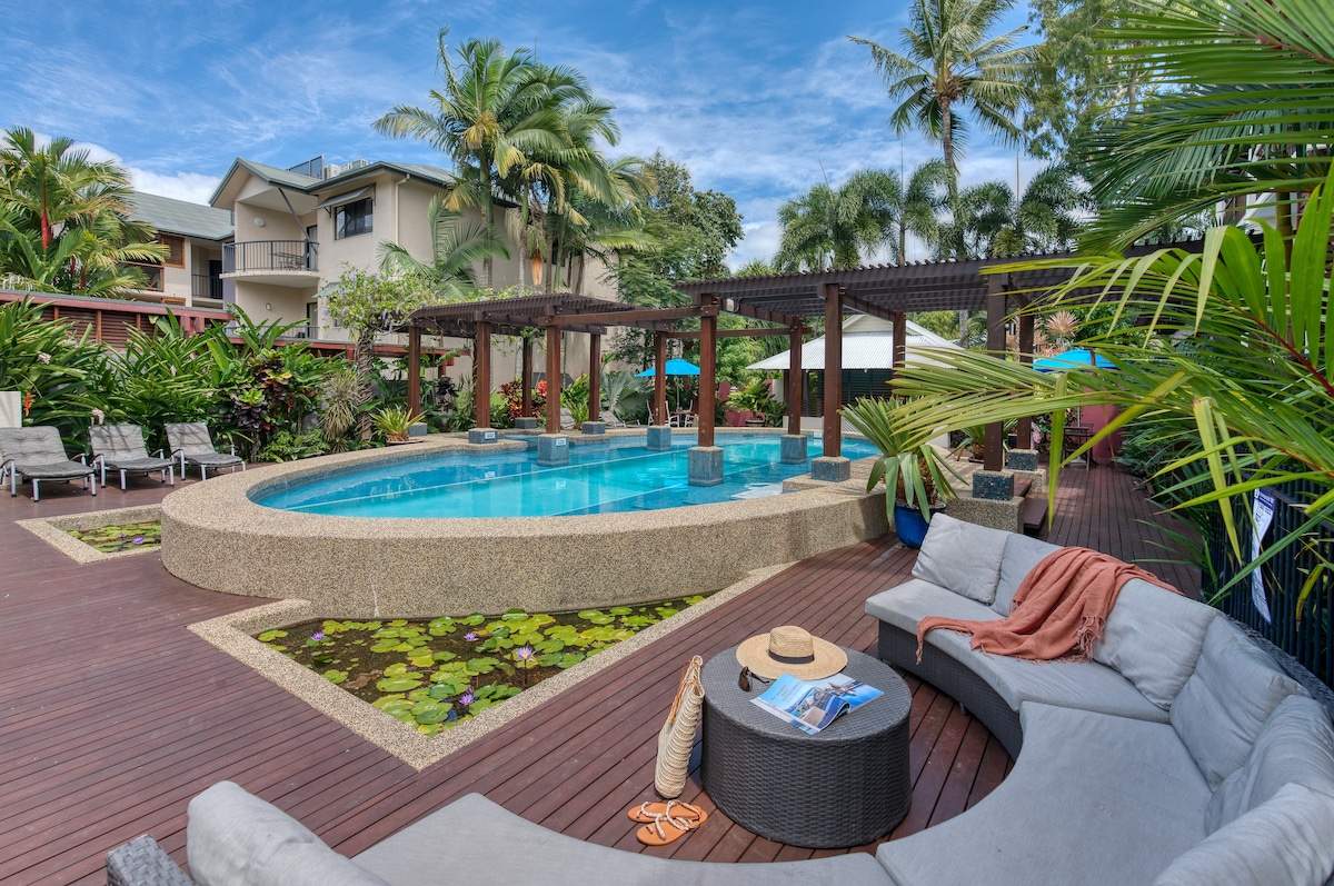 A landscaped pool area is surrounded by tropical foliage and wooden decking. Comfortable lounge chairs are arranged around the pool, with shaded seating nearby. A circular couch is positioned to offer views of the water, creating a relaxing outdoor space for guests.