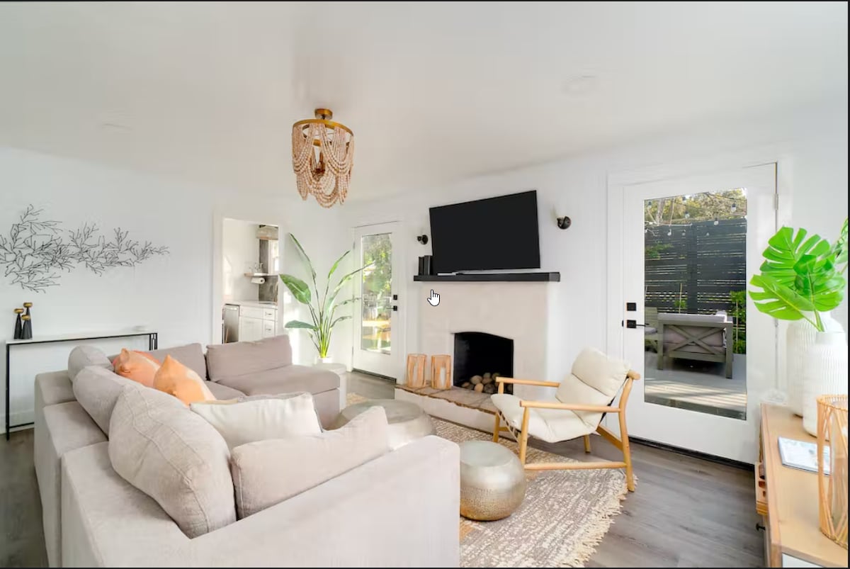 A bright living area is featured with a spacious sectional sofa and an accent chair, beside a modern fireplace. Large windows allow natural light, and a flat-screen TV is mounted above the fireplace. A sliding door leads to an outdoor space, adding connectivity.