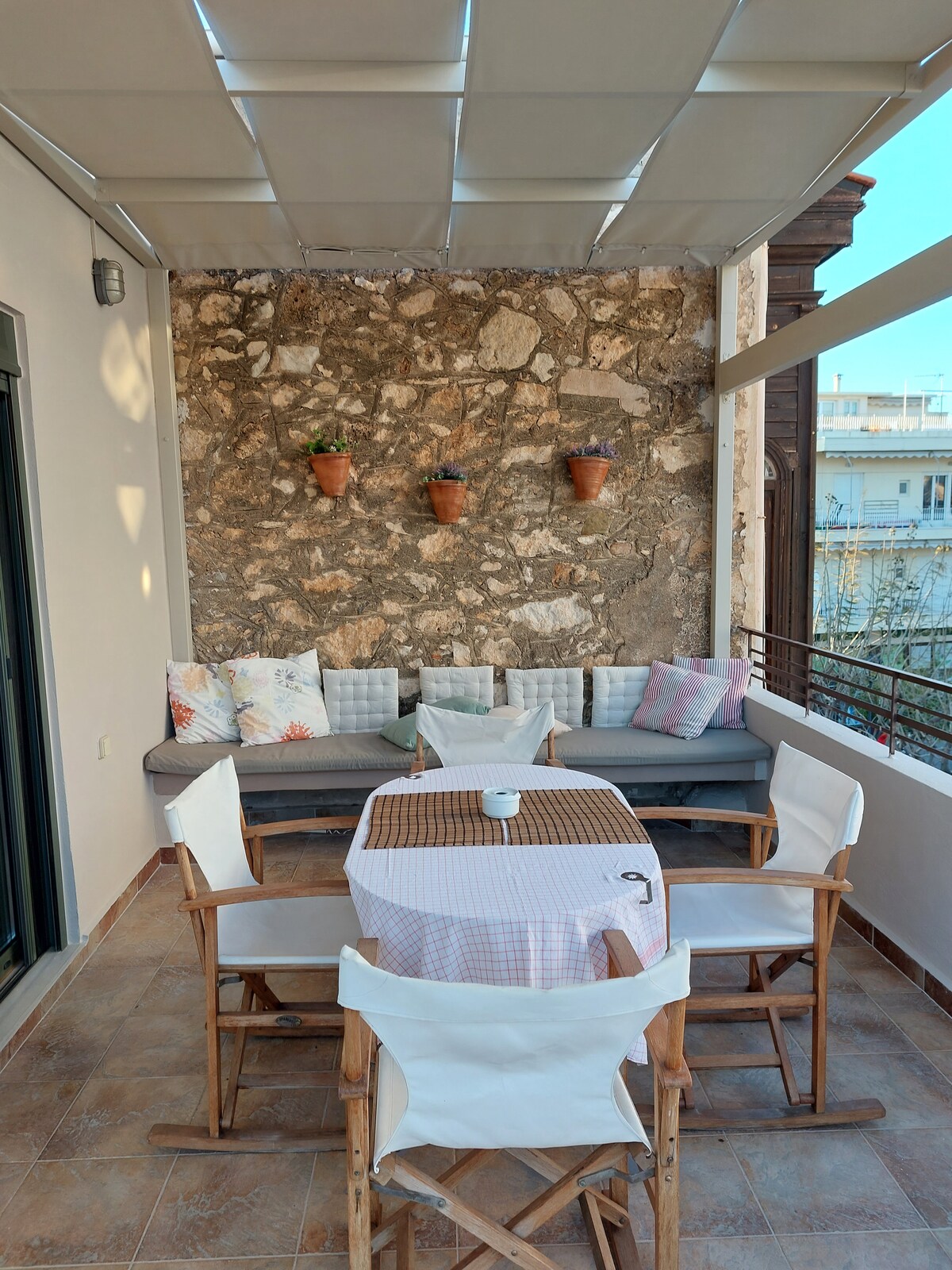 A spacious terrace features a comfortable seating area with soft cushions against a textured stone wall. A dining table surrounded by wooden chairs is positioned nearby. Potted plants add a touch of greenery, enhancing the outdoor atmosphere.