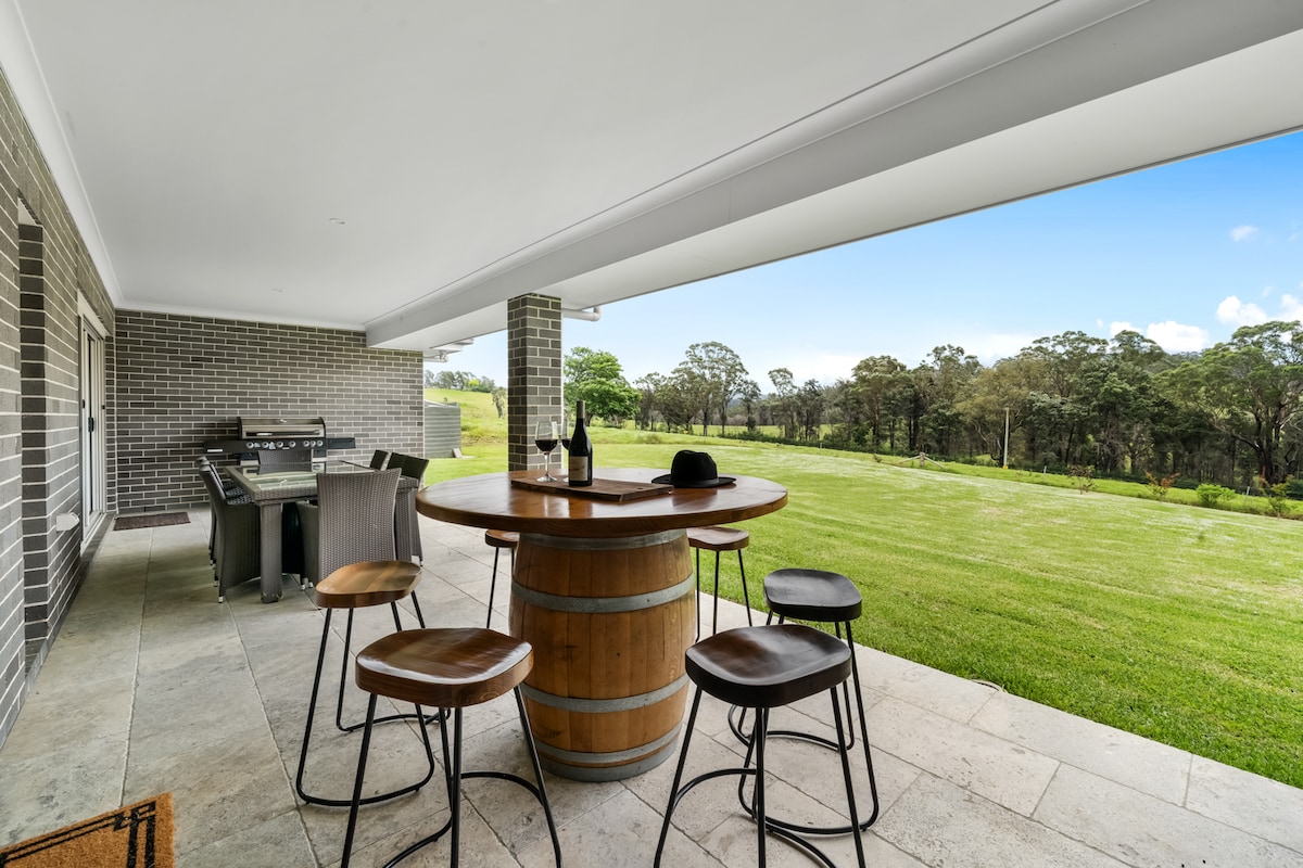 An outdoor patio features a wooden barrel table surrounded by four metal stools. A barbecue grill is positioned nearby, under the cover of the roof. The space overlooks a vast green lawn and a natural landscape, providing a serene outdoor setting.