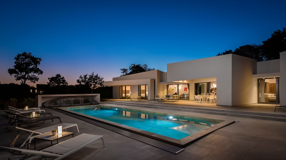 The modern villa is illuminated in the evening, showcasing a heated pool surrounded by lounge chairs. Soft lighting highlights the outdoor living area, which connects to the indoor space through expansive glass doors, creating a seamless blend of comfort and style.