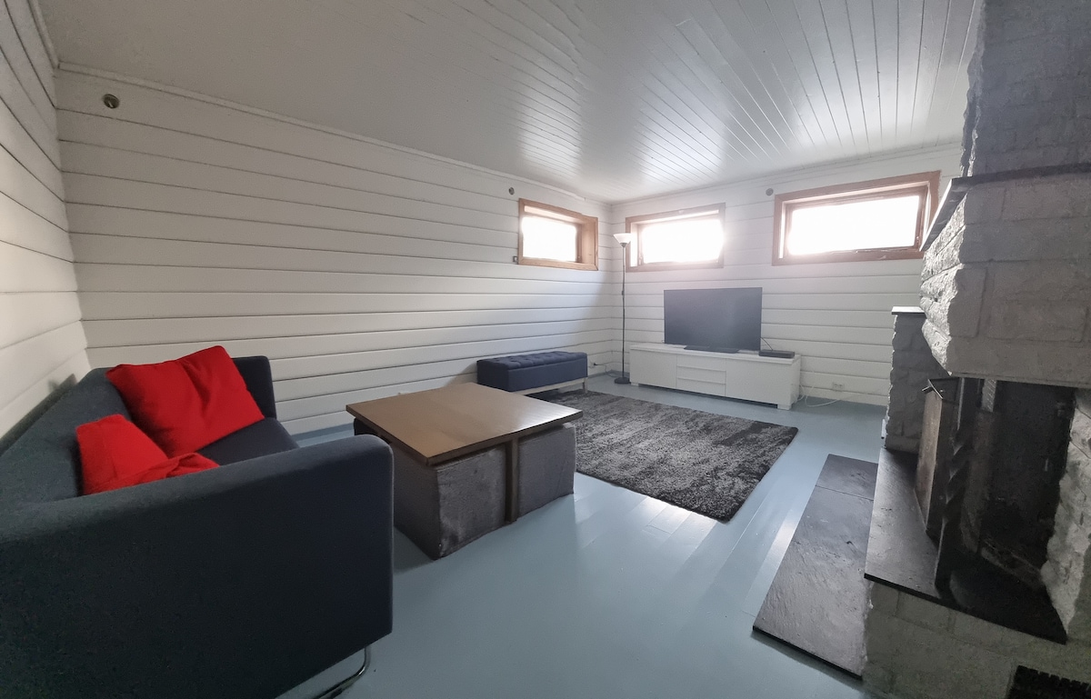 A spacious living area is featured, characterized by a light-colored wooden ceiling and white walls. A large flat-screen television is positioned on a media console. Comfortable seating is provided by a grey sofa adorned with red cushions, paired with a low coffee table. A stone fireplace adds a cozy element to the space.