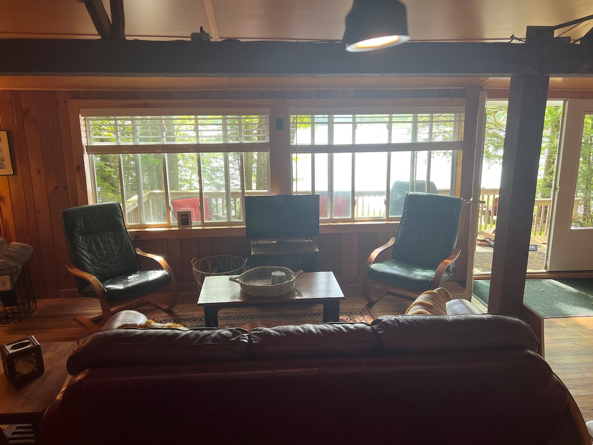 A cozy living area features a leather sofa facing a wooden coffee table. Two green chairs are positioned next to large windows that allow natural light and views of the lake. A television is visible across the room, enhancing the space's relaxation options.