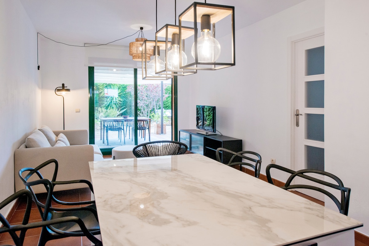 A modern dining area features a spacious marble table surrounded by black chairs. Large glass doors provide access to a private garden. The setting includes a comfortable seating area and a television, enhancing the open and inviting atmosphere.
