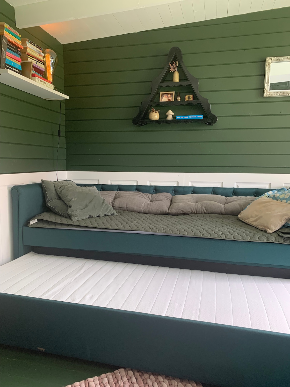 A cozy seating area features a cushioned daybed with a quilted cover, accompanied by decorative pillows. Above, a black shelf displays various decorative items, while the green walls provide a soothing backdrop. A second pull-out mattress is positioned below the daybed.