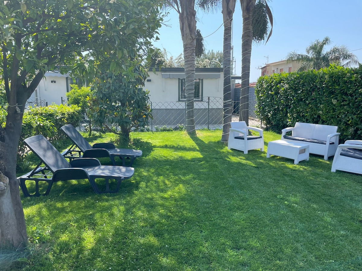 A well-maintained garden area features green grass and palm trees, providing a relaxing setting. Two lounge chairs and a white seating set invite guests to enjoy the outdoor space, which is surrounded by lush greenery and a fence for privacy.