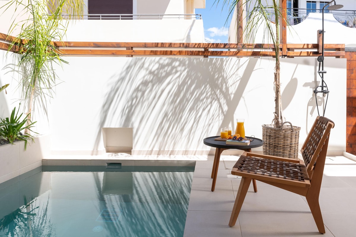 A private outdoor patio features a tranquil heated swimming pool surrounded by sleek, modern tiles. A wooden chair with a woven design sits beside a small table, which holds two drinks. Shadows from palm leaves create a calming visual effect over the space.