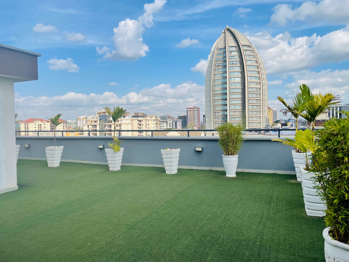 An expansive rooftop terrace features artificial grass and decorative planters, providing a serene outdoor space. The skyline is visible in the background, showcasing a modern building with a distinctive curved design against a clear blue sky.