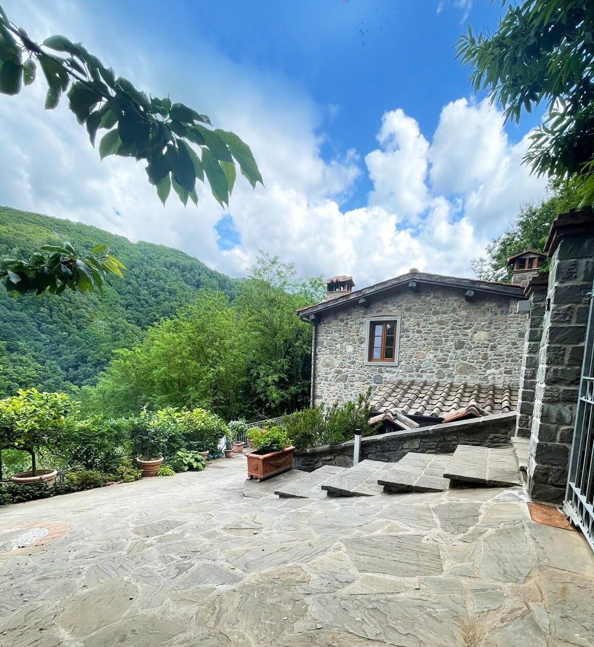 A charming stone house is situated on a sloped pathway, surrounded by lush greenery and hills. Potted plants line the path, and the exterior features stone walls and a tiled roof. A window adds a touch of character, reflecting natural light.
