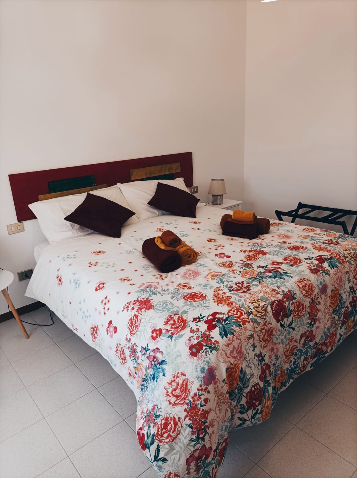 A cozy bedroom features a spacious bed adorned with a floral-patterned blanket and decorative pillows. Two bedside lamps are placed on either side, and a small table sits nearby. Rolled towels are neatly arranged atop the bed, creating a welcoming atmosphere.