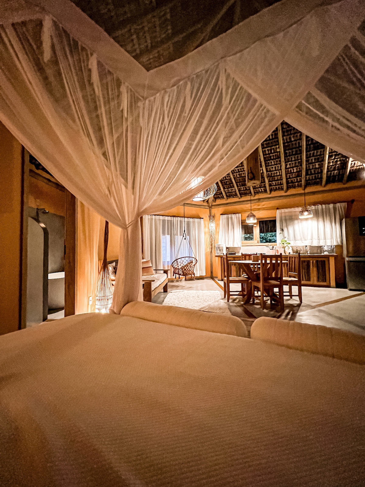 A cozy interior features a spacious bed surrounded by flowing white drapes. A wooden dining area is visible, along with a kitchen in the background. Soft lighting illuminates the space, enhancing the natural textures of the woven ceiling and wooden accents.