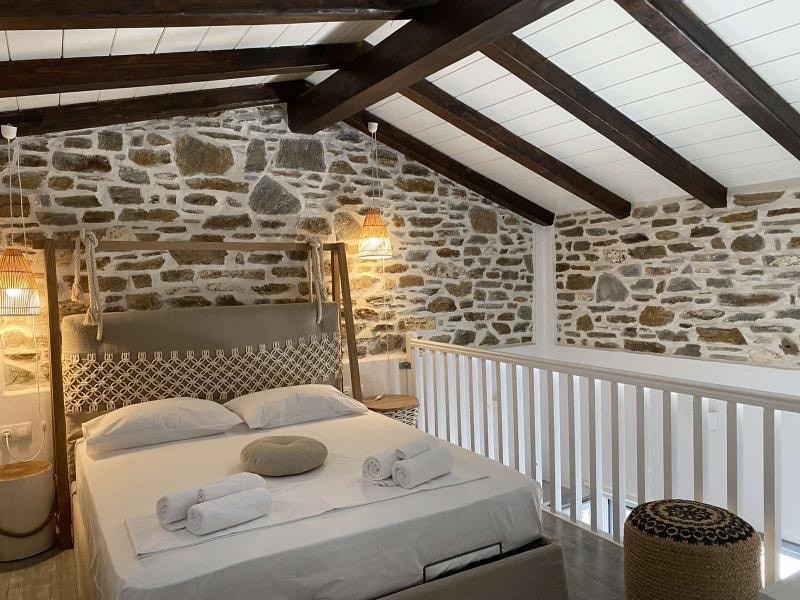 A modern bedroom features a spacious bed with lightly folded towels placed on top. The stone wall provides a rustic backdrop, while wooden beams highlight the ceiling. Warm light is emitted from decorative fixtures, enhancing the cozy ambiance of the space.
