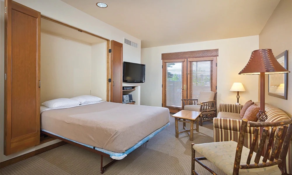 A compact room features a fold-out bed, with tailored bedding and soft pillows, offering a comfortable sleeping area. A small table sits between two seating options, which include a striped lounge chair and a wooden armchair. Natural light filters through the window adorned with wooden blinds.