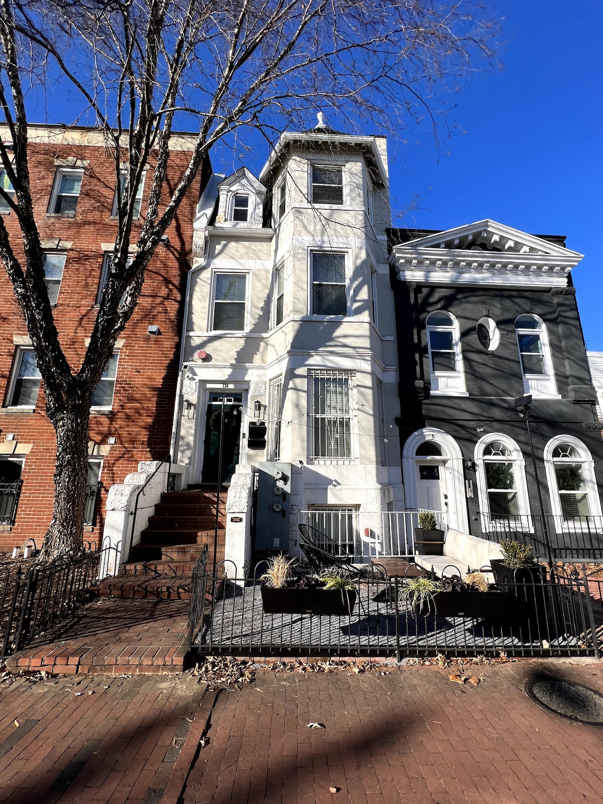 A charming two-story townhouse is displayed, featuring a light-colored façade accented by tall windows and a turret on the roof. A small, fenced front yard with a low brick wall and planter boxes adds character to the welcoming exterior.