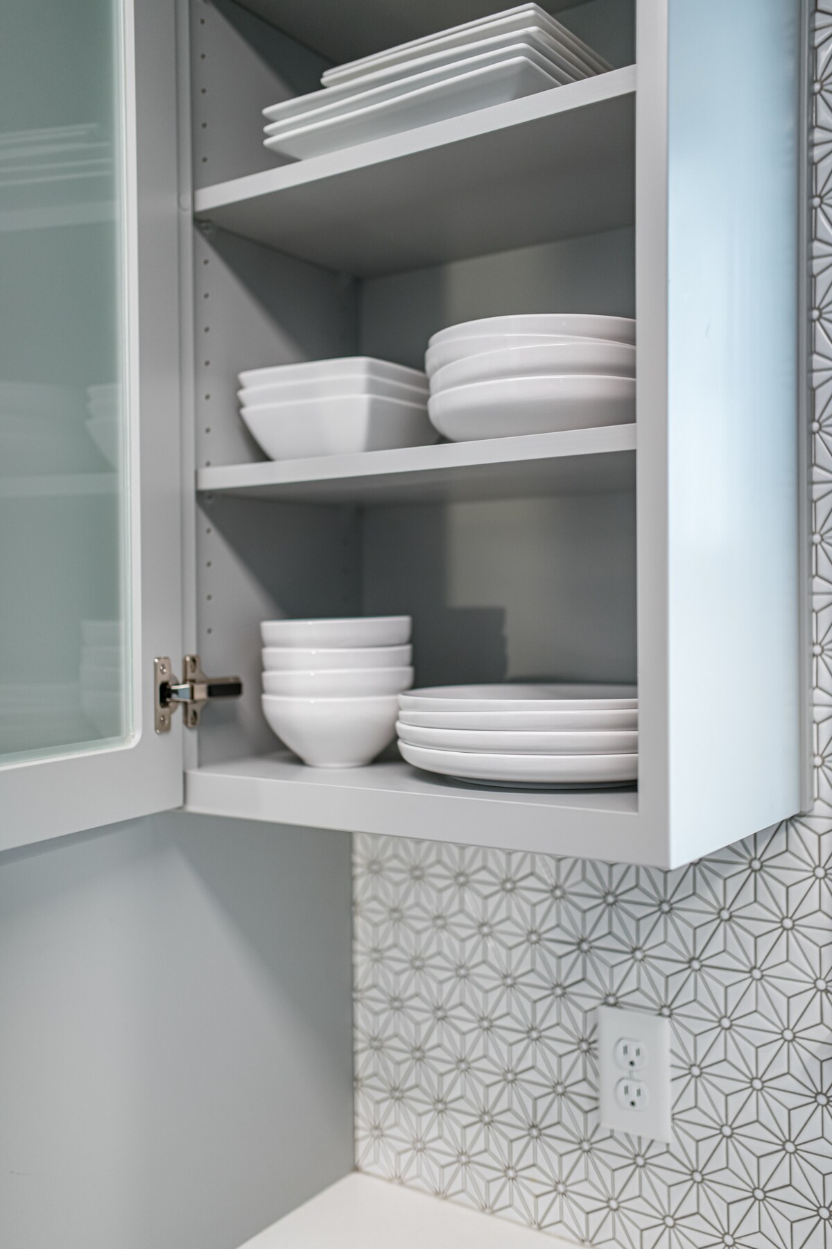 A neatly organized kitchen cabinet displays an array of white dinnerware, including flat plates and deep bowls. The light-colored cabinet contrasts with a patterned wall, contributing to a clean and modern aesthetic.