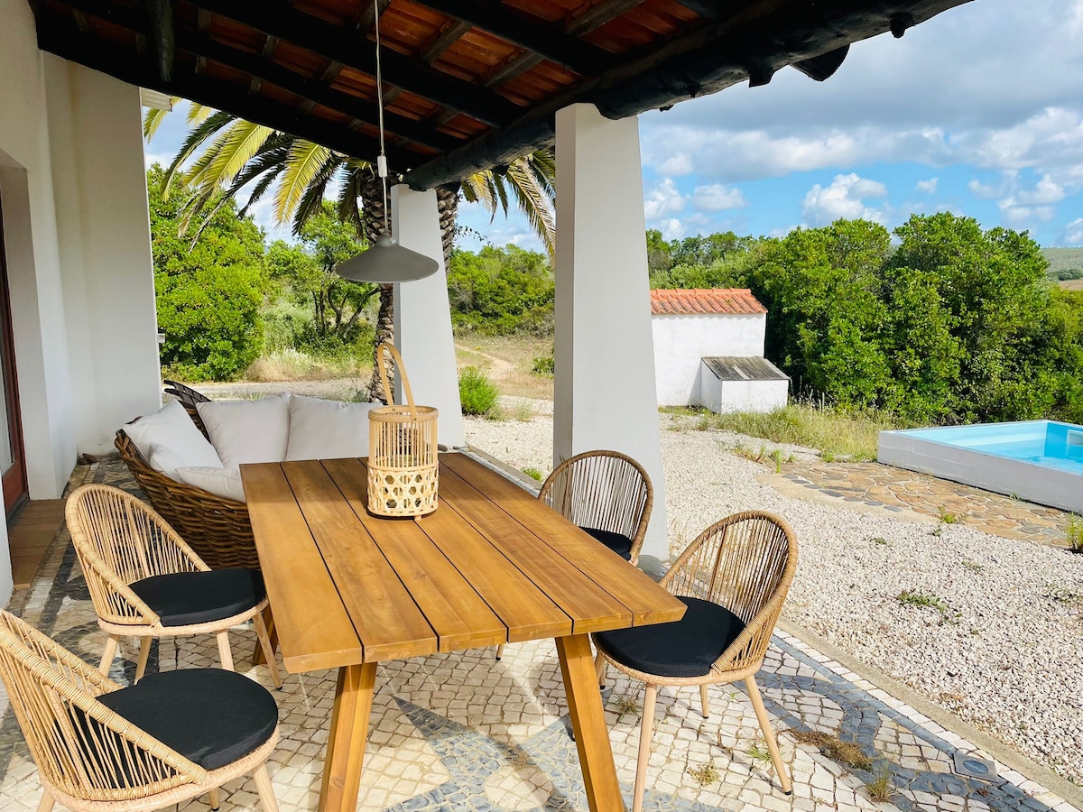 An outdoor terrace is shown with a wooden dining table surrounded by four rattan chairs, each featuring a black cushion. A lantern is placed centrally on the table. Lush greenery and a swimming pool are visible in the background, framed by tall palm trees.