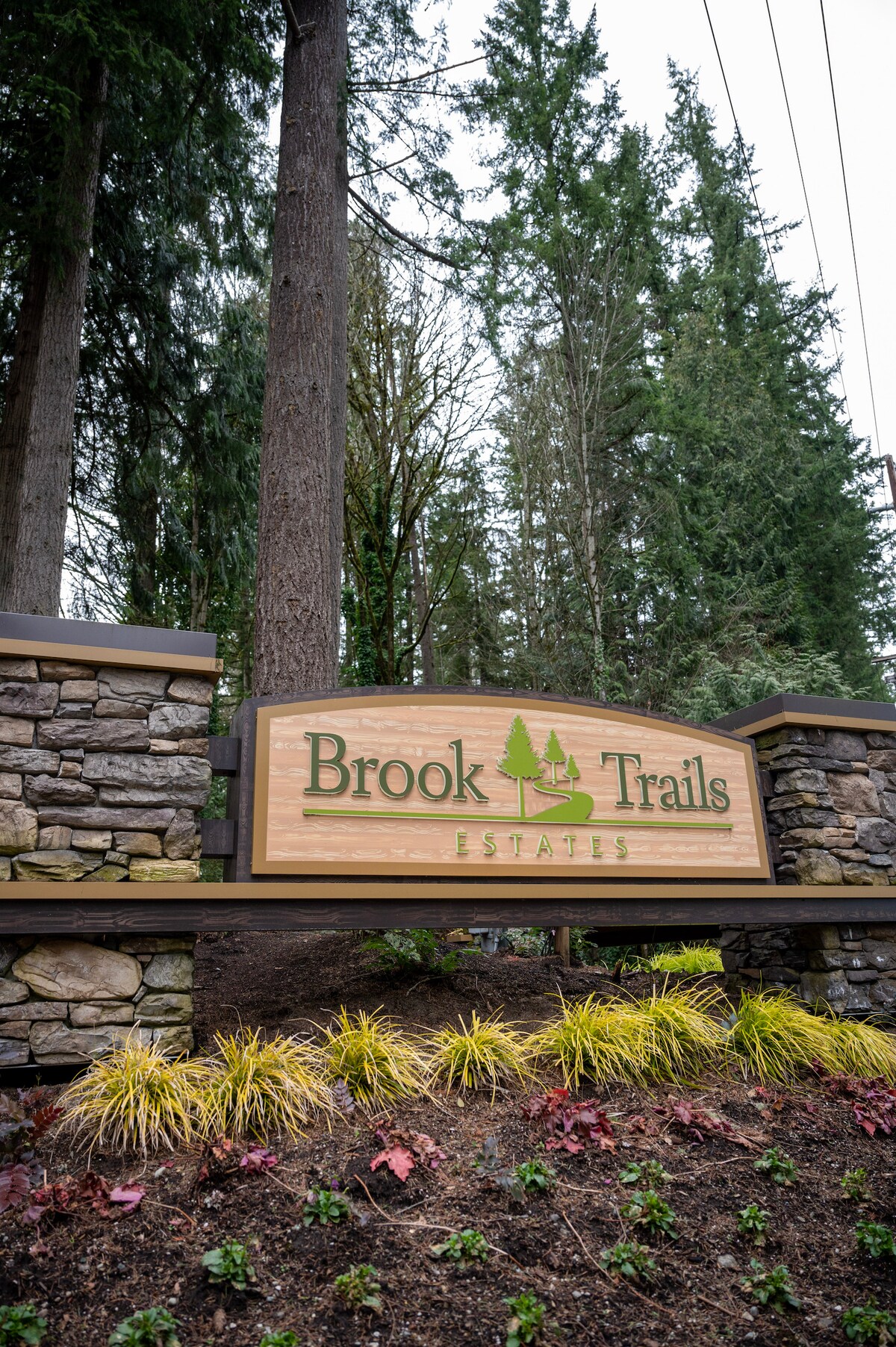 A welcoming entrance sign for Brook Trails Estates is prominently displayed, featuring natural wood and stone elements. Surrounding the sign are landscaped flower beds with evergreen plants and colorful foliage, providing a connection to the surrounding trees and natural setting.