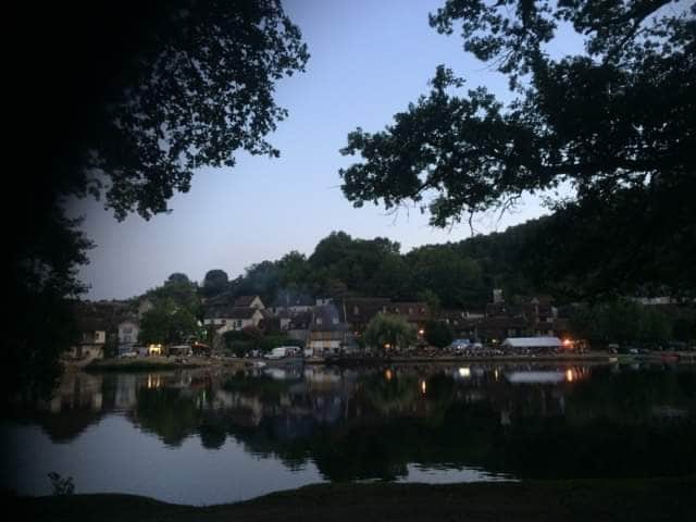 A calm river scene is depicted during twilight, with reflections of quaint houses along the shoreline visible on the water's surface. Trees frame the image, adding depth to the peaceful landscape. Soft lighting from the buildings enhances the tranquil atmosphere.